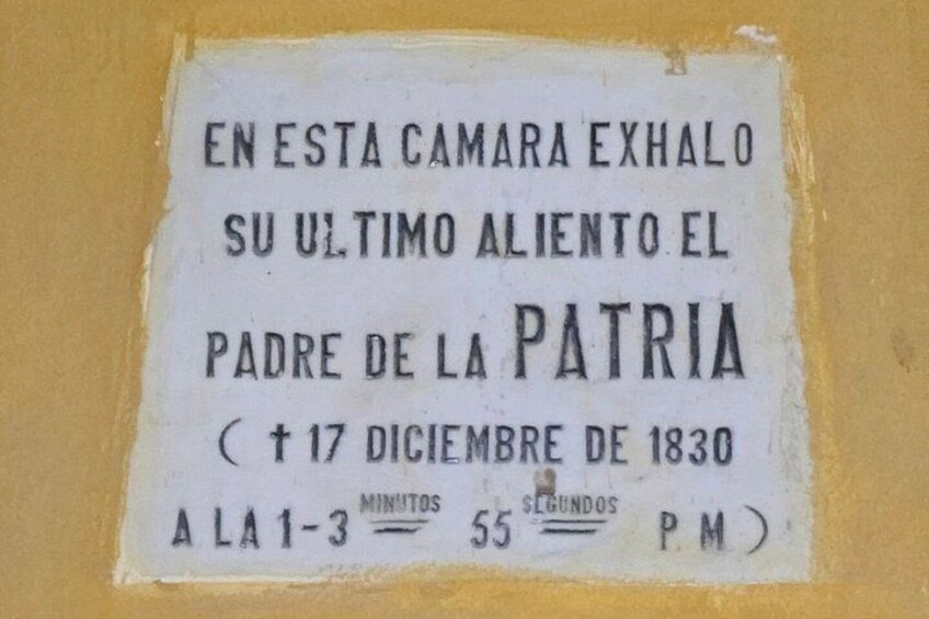 A historical plaque on a yellow wall with text in Spanish, mentioning a significant event on December 17, 1830, and times for a service. 3