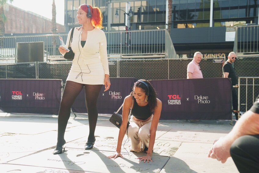 A woman in a white outfit and another in a black top kneeling on a sidewalk with a sign in the background. 2