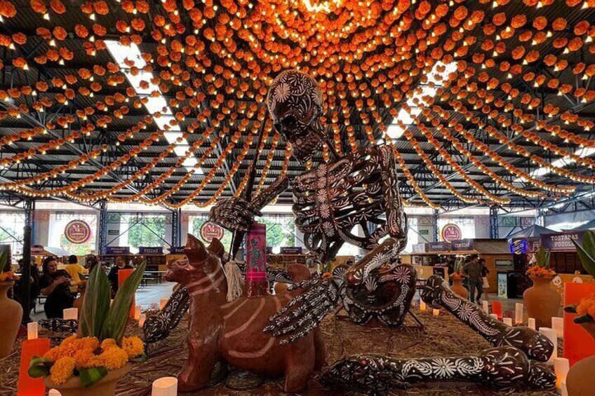 Una gran escultura de esqueleto ricamente decorada en un ambiente festivo, con luces colgantes y arreglos florales. 2