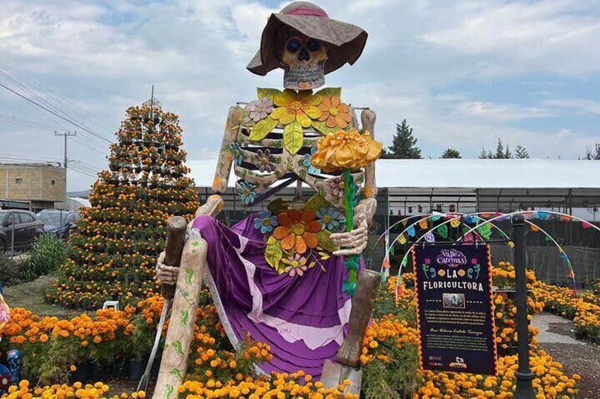 Una estatua de esqueleto sosteniendo flores, rodeada por un jardín de flores y un letrero que dice Florigultora. 4