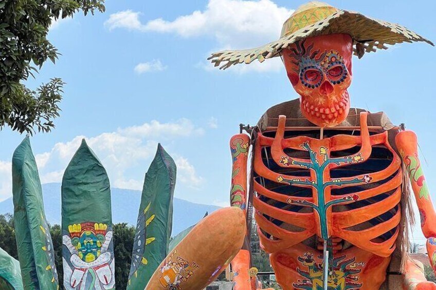 Una figura de esqueleto de gran tamaño vestido con un sombrero, adornado con decoraciones coloridas y un fondo de banderas verdes. 1