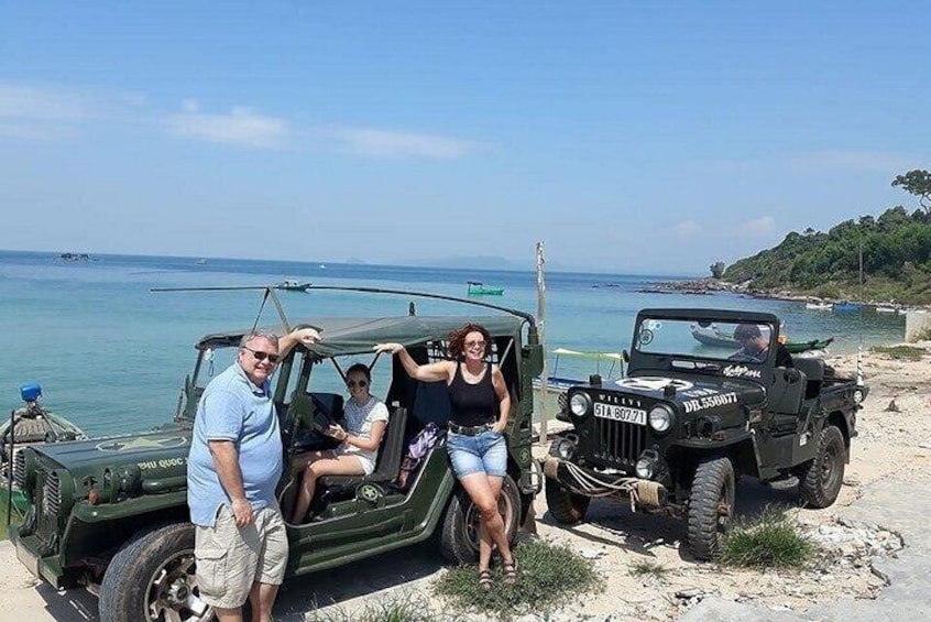 A beach scene with a green jeep and a black jeep parked on the sand, a man standing next to the green jeep, and a woman sitting in the driver's seat of the green jeep. 1