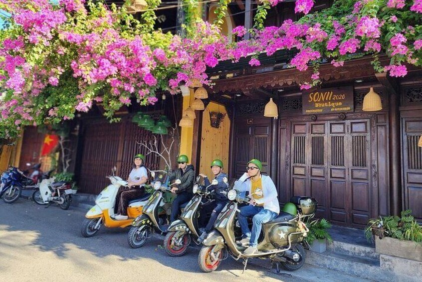 A group of people on scooters parked in front of a building with a sign that reads "SONI 2020". 1