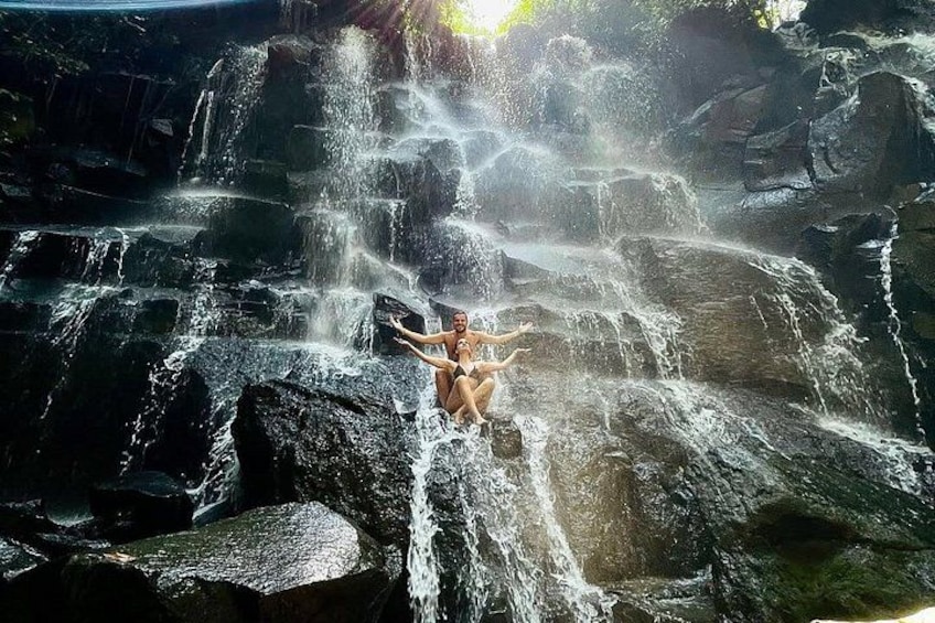 A waterfall with water cascading over rocks, a person sitting on a rock, and sunlight filtering through the trees. 5