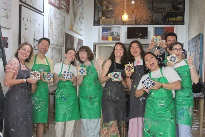 A group of people in a workshop with paint splatters on aprons and walls, holding colorful tiles. 1