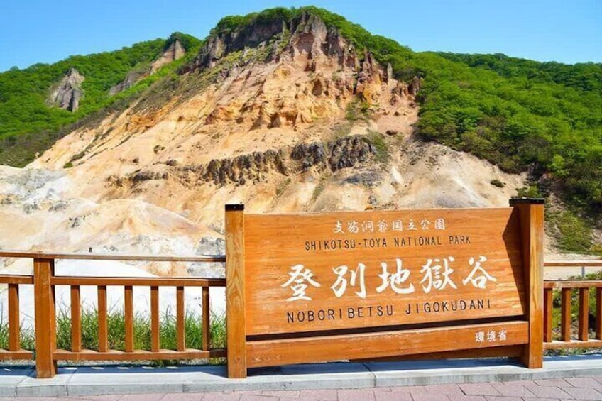 A wooden sign with text in both English and another language, set against a backdrop of a mountainous landscape with greenery. 1