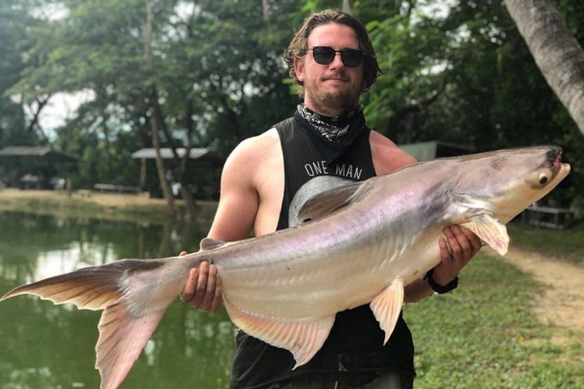 A person holding a large fish by a body of water with trees and a path in the background. 1
