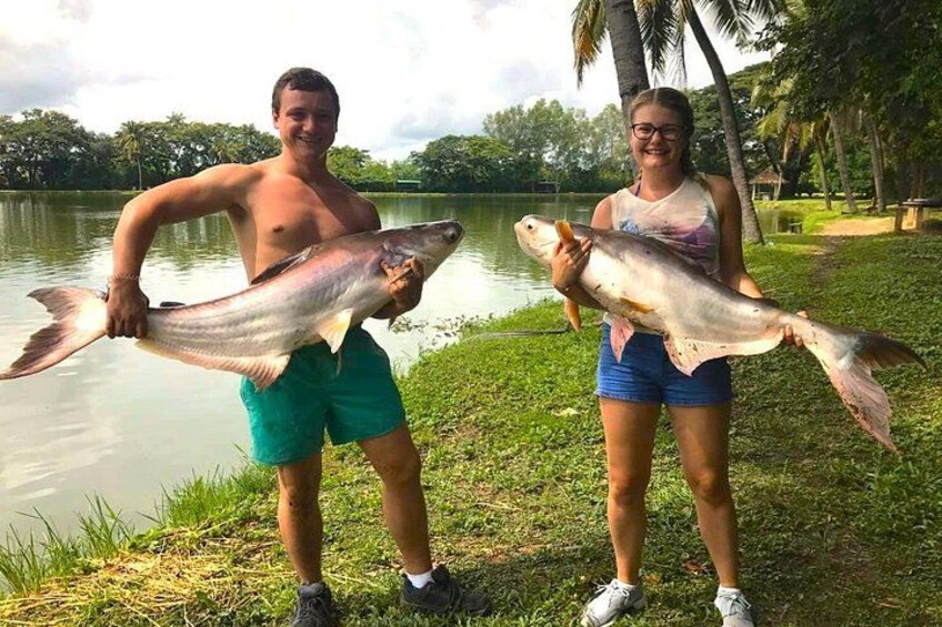 Two people holding large fish by a lake with palm trees in the background. 2