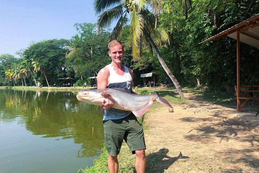 A person holding a large fish by a lake with palm trees and a wooden structure in the background. 3