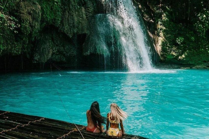 A waterfall in a turquoise pool with two people sitting on a dock. 5