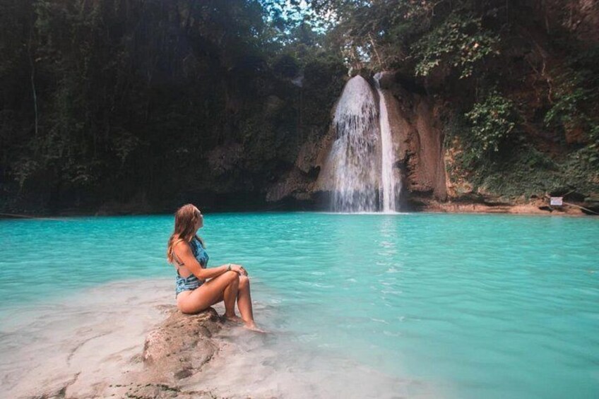 A woman sitting on a rock by a turquoise pool with a waterfall in the background. 3