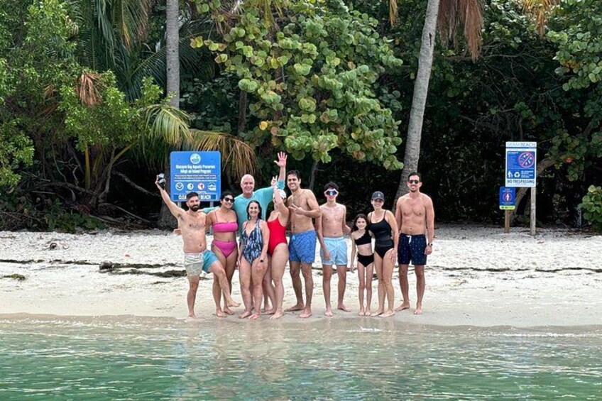 Un groupe de personnes se tient sur une plage, avec des palmiers et un panneau en arrière-plan. 2