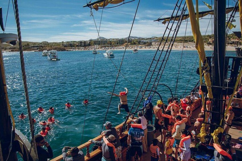 Un bote abarrotado con gente nadando en el agua, una playa y varios botes al fondo. 2