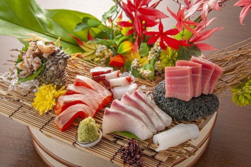 A variety of sushi and sashimi on a bamboo mat, surrounded by decorative elements like flowers and a stone. 1