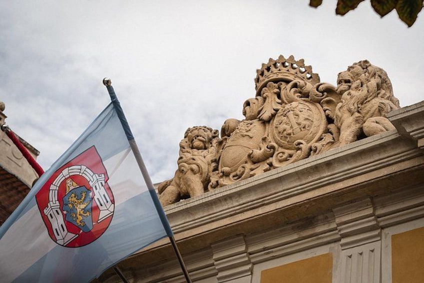 A flag with a coat of arms, a stone lion sculpture, and a building facade. 1