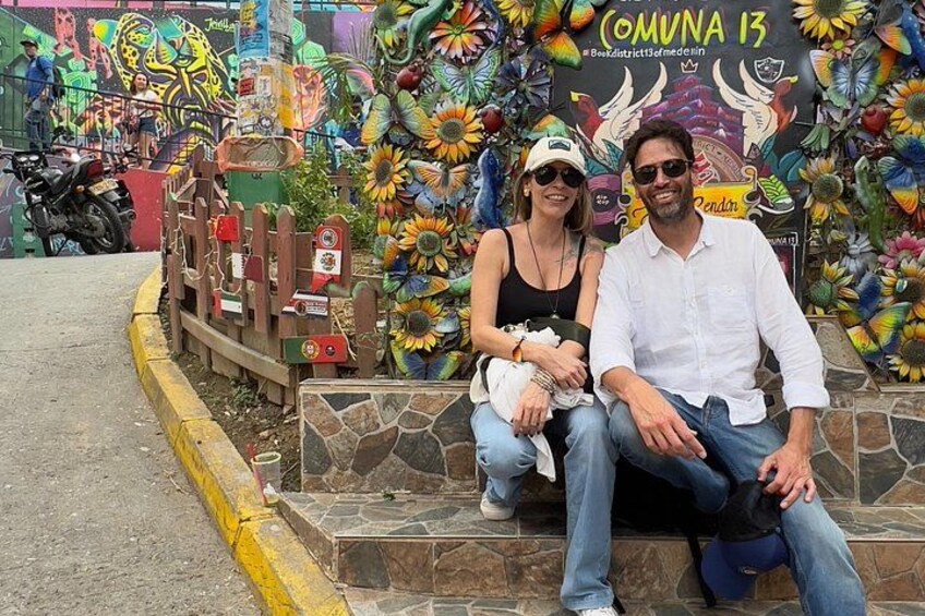 A couple sitting on a stone bench in front of a colorful mural with sunflowers and text. 1