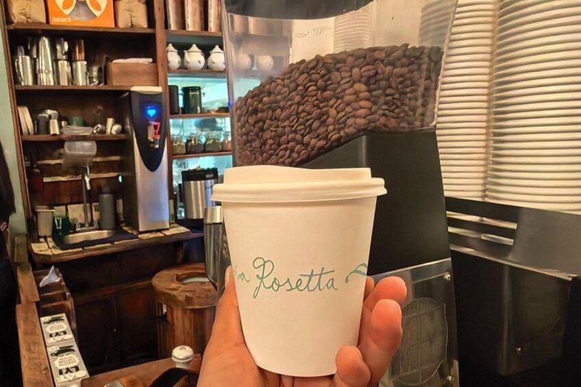 En hand håller i en engångskaffemugg med texten ”la Rosetta” på, i ett kafé där en kaffemaskin och hyllor med köksredskap syns i bakgrunden. 5