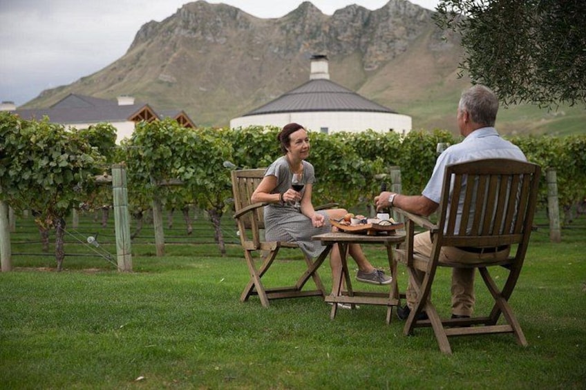 Two people sitting at a wooden table outdoors with a vineyard and mountains in the background. 5