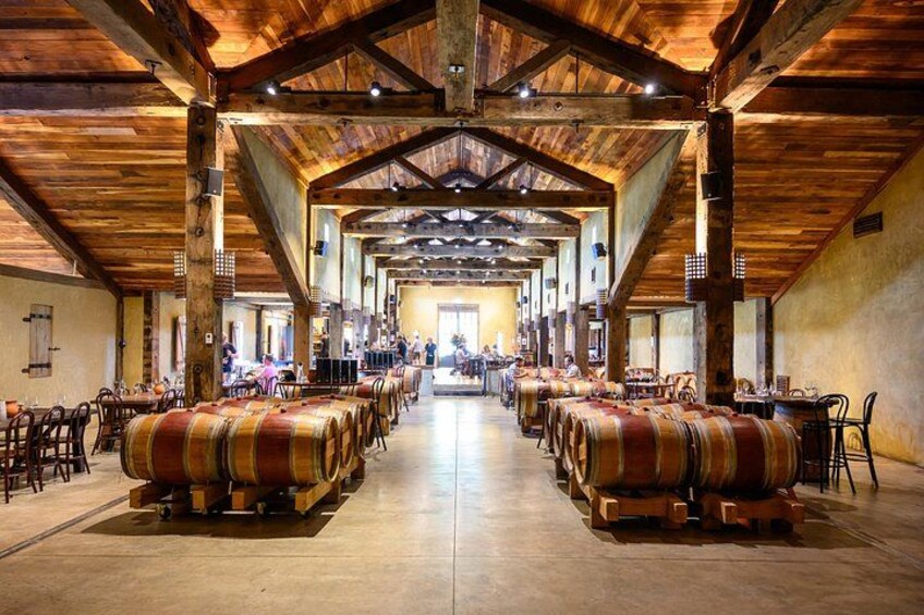 A spacious hall with wooden beams and tables, barrels lined up in the center, and a long table at the far end. 4