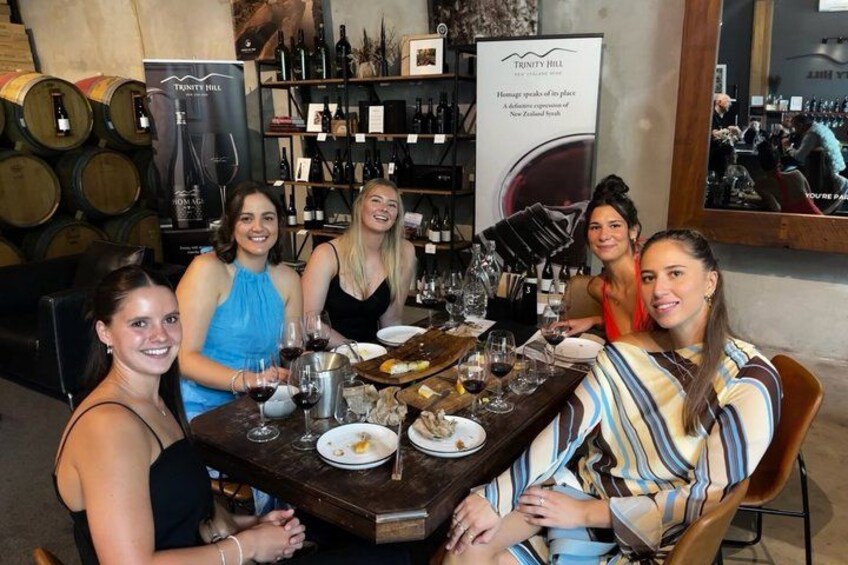 A group of people seated at a table in a wine cellar with wine bottles and a banner in the background. 2