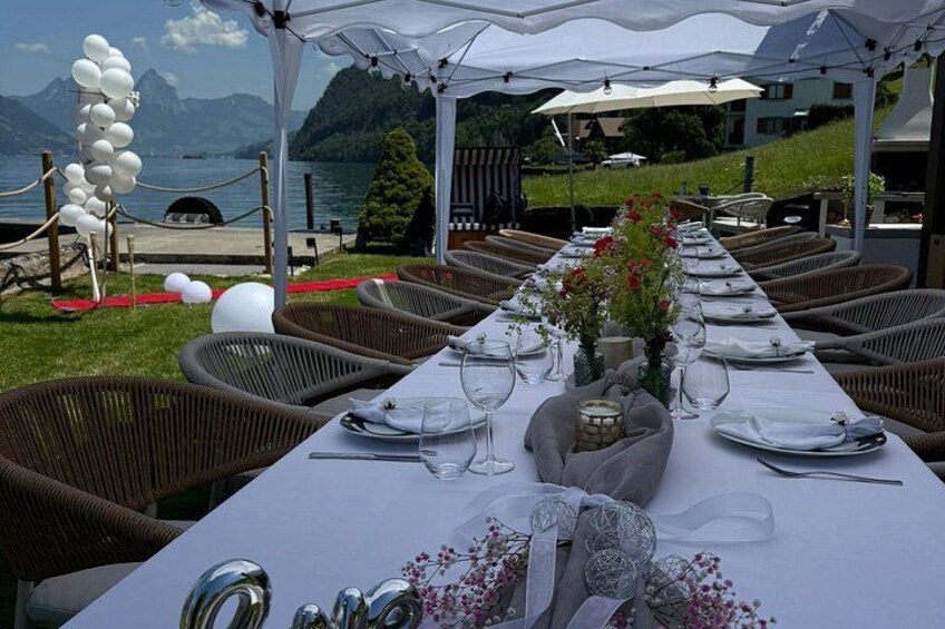A long dining table set for a meal with wicker chairs, white tablecloth, and a scenic lake view. 4