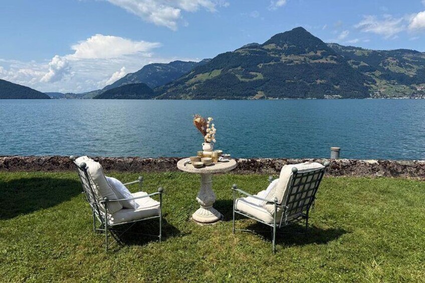 Two white chairs and a table with a vase of flowers on a grassy area by a lake with mountains in the background. 1