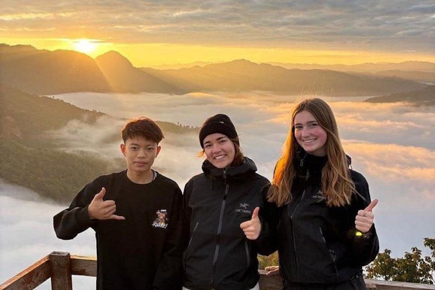 Drie personen geven een duimpje omhoog voor een bergachtig landschap bij zonsopgang. 1