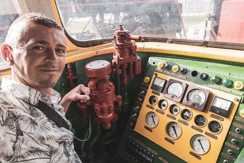 A man in a patterned shirt standing in front of a control panel with gauges and red valves. 3