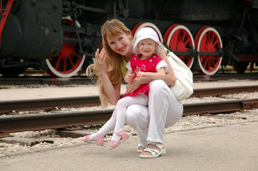 A woman and a child sitting on the ground near train tracks with a steam locomotive in the background. 1