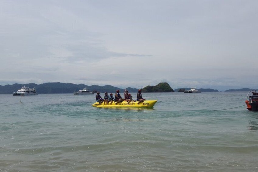 A yellow boat with people on a calm sea, surrounded by larger boats and distant islands. 4