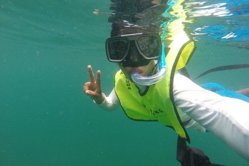 En person under vatten, med en snorkelmask och en gul flytväst på sig, som gör en fredsgest. 2