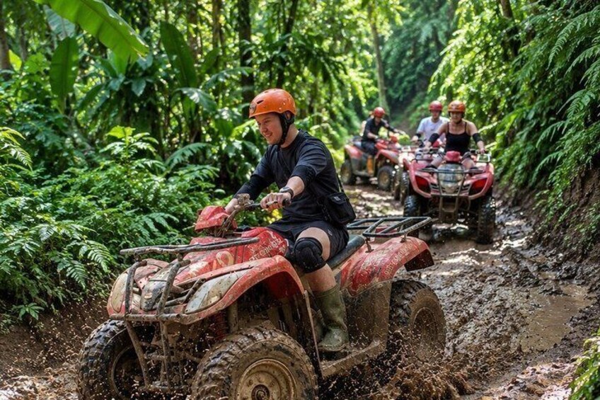 A person riding an ATV through a muddy path surrounded by dense green foliage. 1