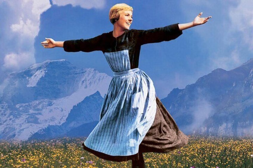Una persona con vestido azul, brazos extendidos, en un campo de flores amarillas, con montañas al fondo. 1