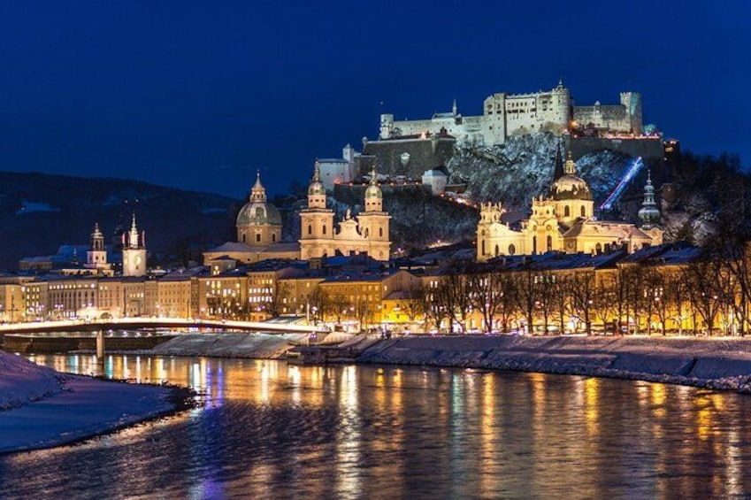 Eine historische Stadtansicht mit beleuchteten Gebäuden, einem Fluss und einer Burg auf einem Hügel. 2