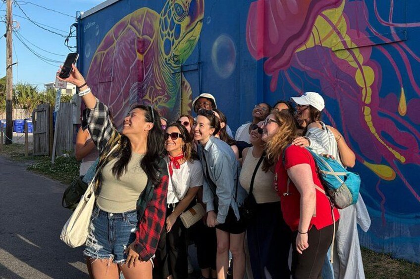 A group of people taking a selfie in front of a colorful mural. 1