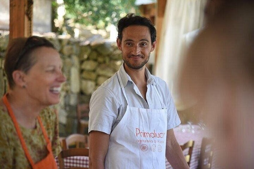 A person wearing a white apron with text, standing in an outdoor setting with stone walls and greenery. 2