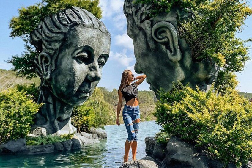 A person standing near a large sculpture of a face in a natural setting with water and greenery. 1