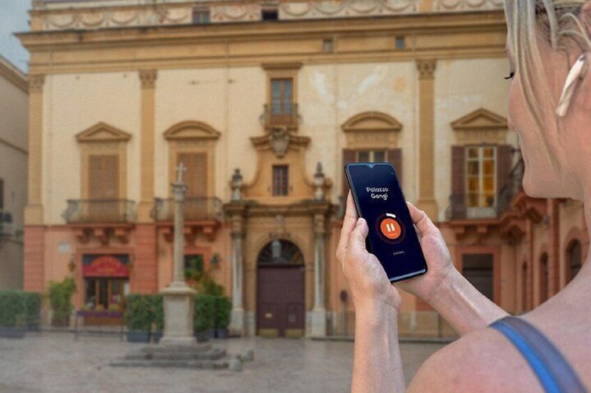 A person holding a smartphone with a game app open, standing in front of a historic building with a fountain and a red awning. 4
