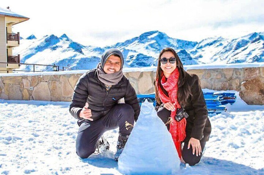 Dos personas posando junto a una escultura de nieve en un paisaje nevado con montañas al fondo. 1
