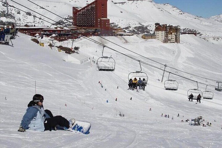 Una pista de esquí nevada con una persona sentada sobre una tabla de snowboard, telesillas y edificios al fondo. 3