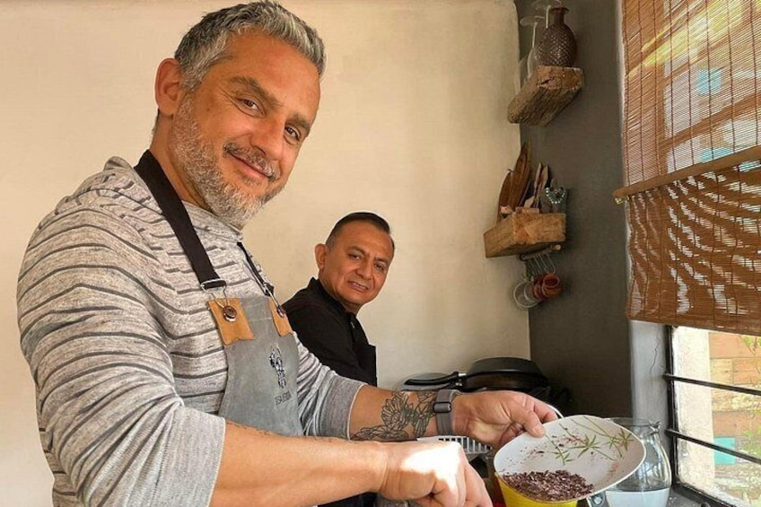 A person cooking with a pot on a stove, another person in the background, and kitchen utensils on the wall. 1