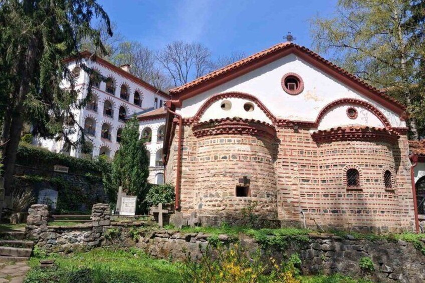 Eine historische Kirche mit Ziegelverkleidung und weißen Wänden, umgeben von Grünpflanzen und einer Steinmauer. 5