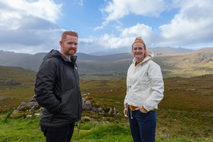 Two people standing on a grassy hill with a mountainous landscape in the background. 2