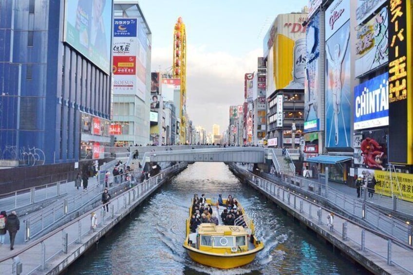 Un bote amarillo con pasajeros en un canal en una zona urbana concurrida, con edificios altos y publicidad. 2