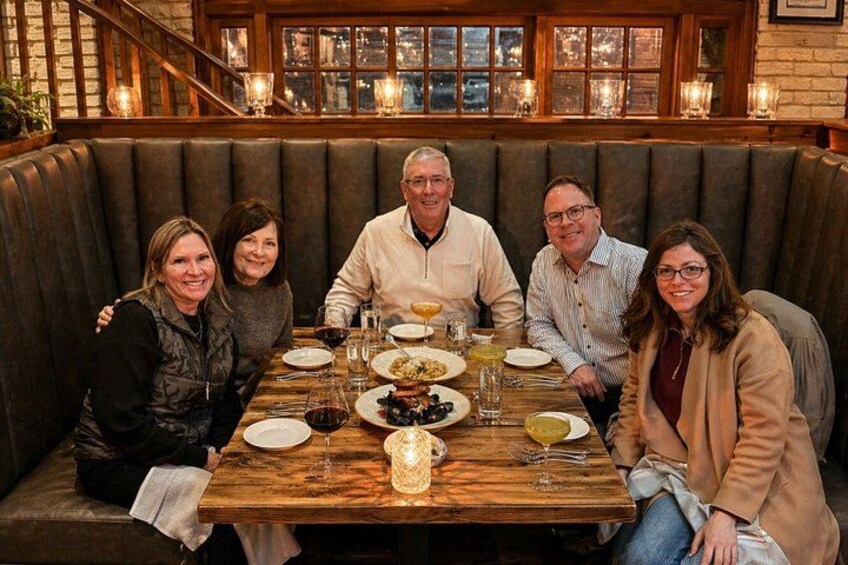 A group of people seated at a wooden table in a restaurant with a brick wall and windows in the background. 1