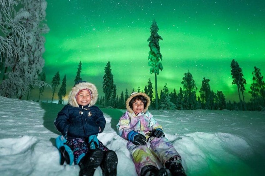 To personer sidder i sneen med et grønt nordlys i baggrunden. 3