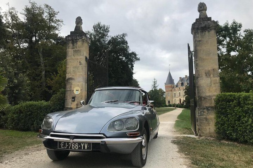 A classic car parked on a gravel path with stone pillars and a gate, leading to a historic building. 2