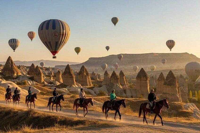 Un grupo de personas montando a caballo por un sendero de tierra con globos aerostáticos al fondo. 2