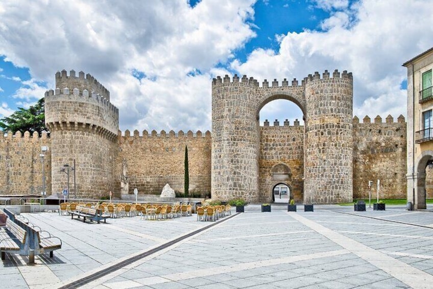 A historic stone gate with a central archway, flanked by two round towers, in a paved courtyard with benches. 5