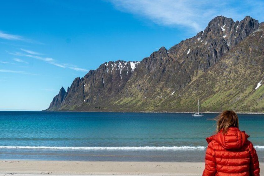 Une personne vêtue d’une veste rouge se tient sur une plage, face à une chaîne de montagnes, et on aperçoit un voilier sur l’eau. 4
